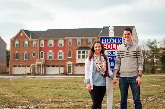 A young couple stands smiling in front of a "Home Sold" sign, symbolizing the beginning of their new chapter. Casually dressed, they stand on a grassy area with newly built townhouses in the background. The day is overcast, and the couple appears happy with their new purchase.