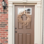 A front porch with a brown door adorned with a rustic wreath and white trim. Below, a white pumpkin rests on the left, while colorful chrysanthemums sit on the right atop a checkered black and white mat. A brick wall and lantern complete this Modern Fall Front Porch. Text at the top reads, "A Simple & Modern fall front porch.