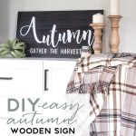 A cozy autumn-themed display featuring a wooden sign with "Autumn - Gather the Harvest" text, two wooden candle holders with white candles, a green succulent, and a plaid blanket draped over a white and black cabinet. Text at the bottom reads, "DIY Easy Autumn Wooden Sign.