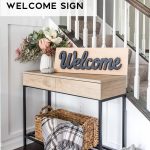 A stylish entryway table displays a DIY Easy Welcome Sign with 3D text. The table holds a vase of flowers, a basket with a cozy plaid blanket draped over it, and a small white pumpkin. A staircase with wooden banisters is seen in the background, perfectly framing the welcoming scene.