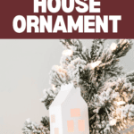 A white 3D paper house ornament with windows glows warmly as it hangs on a snow-dusted Christmas tree branch. The background showcases the text "3D Light-Up House Ornament" alongside the Cricut logo, bringing a modern touch that beautifully complements your merry Christmas décor.