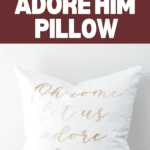 A white pillow beautifully inscribed with "Oh come let us adore him" in gold script rests against a rich burgundy background. At the top, the phrase "OH COME LET US ADORE HIM PILLOW" gracefully highlights its allure, while a convenient website link sits at the bottom.