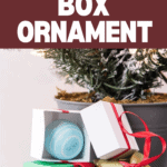 A stack of small, elegantly wrapped boxes with ribbons sits next to an open box unveiling a circular ornament inside. In the background, a Christmas tree twinkles softly. At the top, the text reads "DIY Folding Gift Box Ornament.