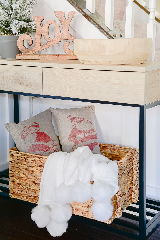 woven basket under console table filled with two Santa pillows and a white pom pom throw blanket