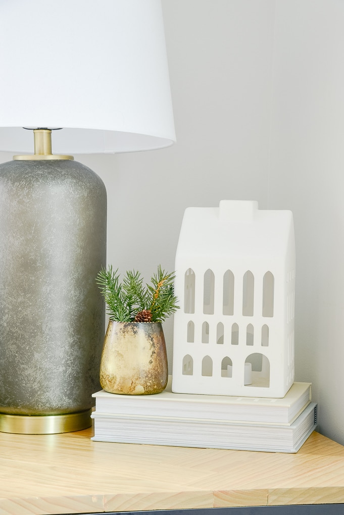 white ceramic Christmas house on end table stack of books and gold vase with greenery next to it