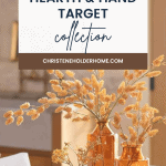 A cozy, fall-themed decor image featuring a wooden table with two amber-colored glass vases filled with dried autumnal foliage. A white banner above the table reads, "Hearth and Hand with Magnolia Target Fall Collection" with a website link "CHRISTENEHOLDERHOME.COM.