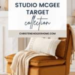 A cozy, stylish living room scene featuring a brown cushioned chair with light wooden legs. The chair is adorned with a beige throw blanket and matching pillow. Behind it is a white cabinet with minimalistic decor. Text overlay reads: "New! Studio McGee Target Spring Collection".