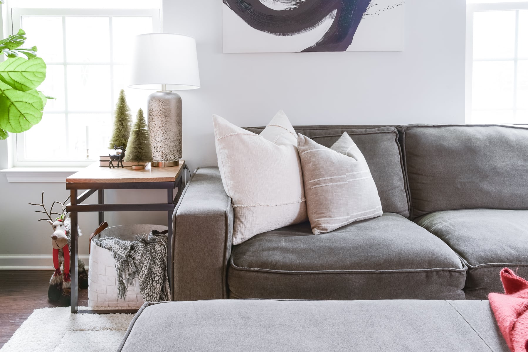 A cozy living room featuring a gray sectional couch adorned with white throw pillows sets the scene for a Christmas Home Tour. A side table holds a lamp, small decorative trees, and reindeer ornaments. The space is brightened by windows with white frames, while a houseplant is visible on the left.