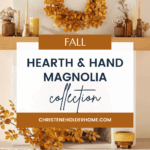 A cozy living room decorated for fall with items from the Hearth and Hand with Magnolia Target Fall Collection. A wreath of autumn leaves hangs above a fireplace mantel adorned with candles and small decor. Below, a table is set with a vase of fall foliage, a cup, and a book.