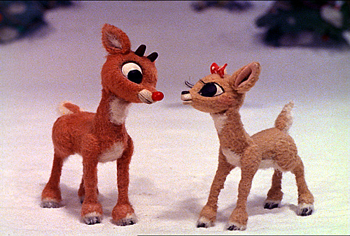 Two stop-motion reindeer characters stand facing each other on a snowy surface. The one on the left has a red nose and the one on the right has a bow on its head. Their wide eyes, open mouths, and animated expressions suggest they are in conversation—a perfect scene from one of the best Christmas movies for 4-year-olds.