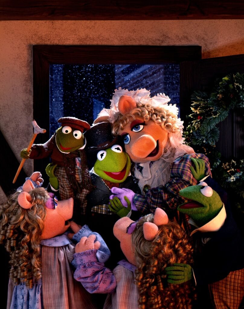 A cozy holiday scene featuring several puppet characters dressed in winter clothes evokes the charm of the best Christmas movies for 4 year olds. Some characters are seen warmly hugging each other while others look on joyfully. There's a wreath on the door, and snow falls outside, enhancing the festive atmosphere.