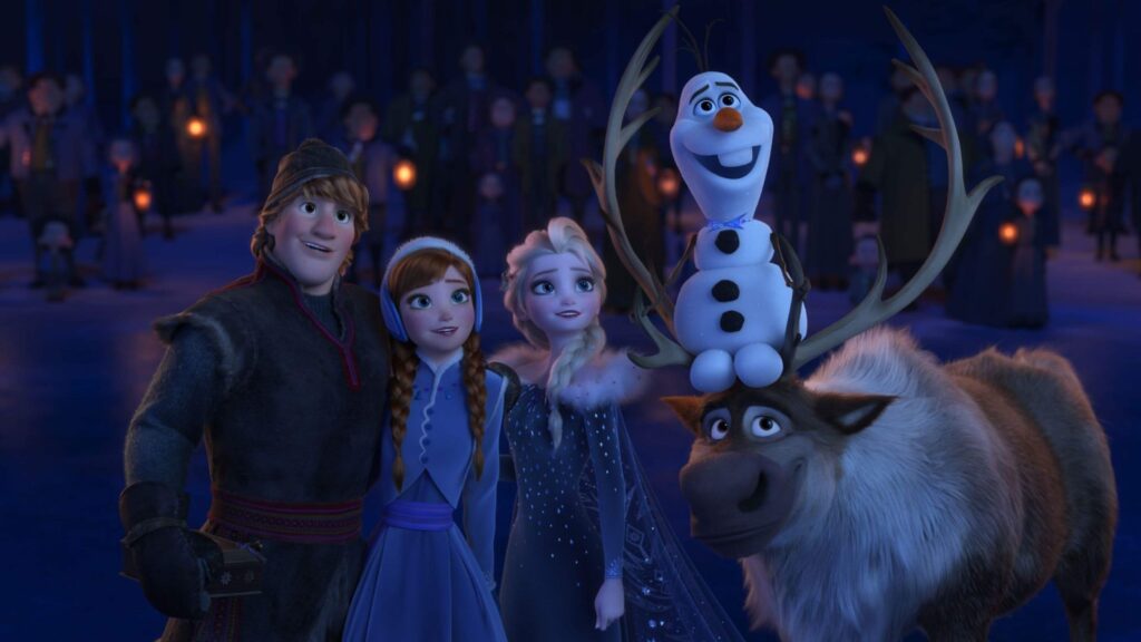 Animated characters Kristoff, Anna, Elsa, Olaf the snowman, and Sven the reindeer are standing together in a snowy forest, smiling and looking upwards. A crowd with lanterns stands in the background, creating a festive, night-time atmosphere straight out of one of the best Christmas movies for 4-year-olds.