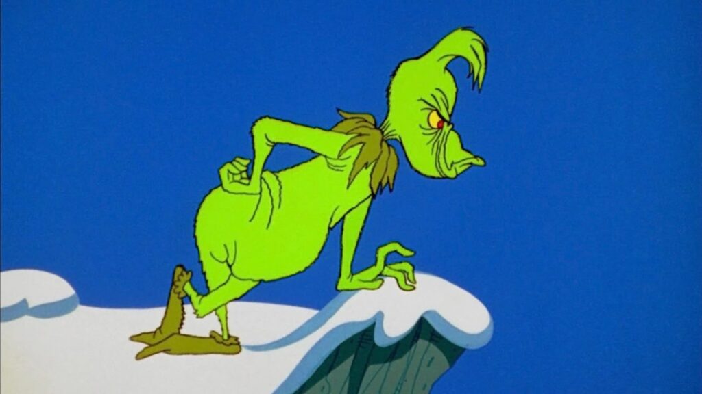 An animated image of the Grinch, a green creature with a tuft of hair on his head, standing hunched over on a snowy cliff edge. The Grinch has a mischievous expression on his face and is looking down into the distance, framed by a clear blue sky; he's from one of the best Christmas movies for 4 year olds.