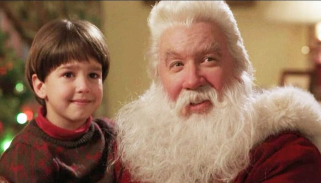 A young boy with dark hair stands smiling next to an elderly man dressed as Santa Claus with a white beard and red suit. They are indoors, festive decorations visible in the background, reminiscent of scenes from the best Christmas movies for 4 year olds.