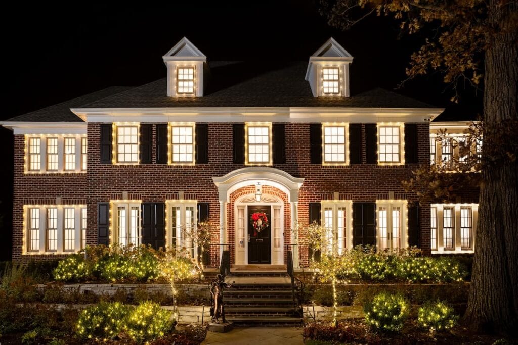 A two-story brick house decorated for the holidays, reminiscent of scenes from the best Christmas movies for 4-year-olds. Symmetrical arrangements of windows outlined with white lights and a main entrance adorned with a red wreath create a magical atmosphere. The pathway and shrubbery are also lit with festive lights.