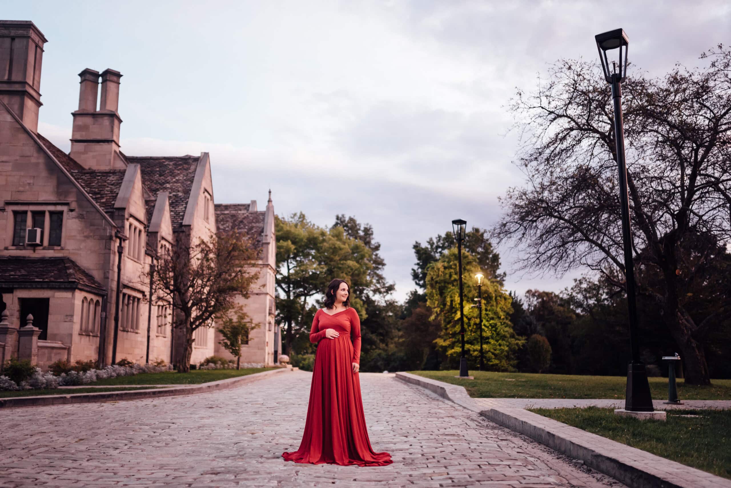 A pregnant woman in a flowing red maternity gown stands on a cobblestone path lined with lampposts. She is positioned in front of a large, stone building with pointed roofs and tall chimneys on a cloudy day, with trees and greenery surrounding the scene.