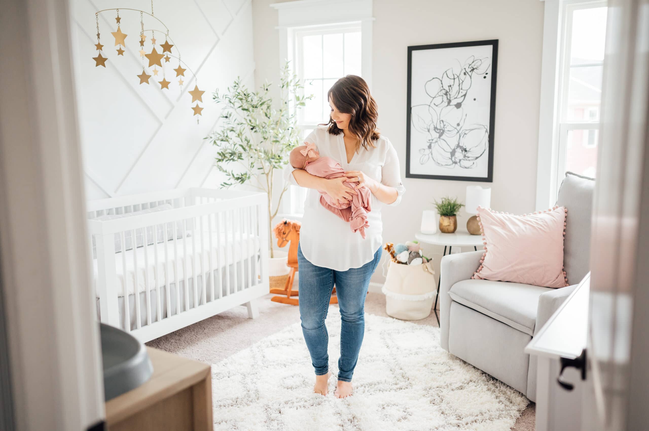 A woman stands in a softly lit nursery holding a baby wrapped in a pink blanket. The room, optimized for a growing family, is decorated with a crib, a plush chair, and various baby items. Star-shaped decorations hang from the ceiling, and a black-and-white art piece adorns the wall.