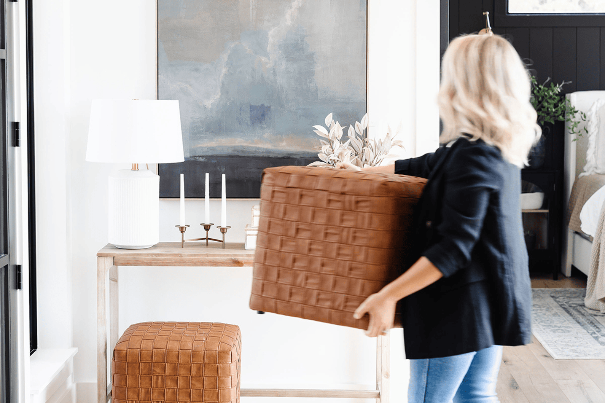 A person with blond hair, wearing a black jacket and jeans, is moving a light brown woven ottoman in a stylish, practical home. The room features a console table with decorative items including a lamp, painting, and candlesticks in the background.