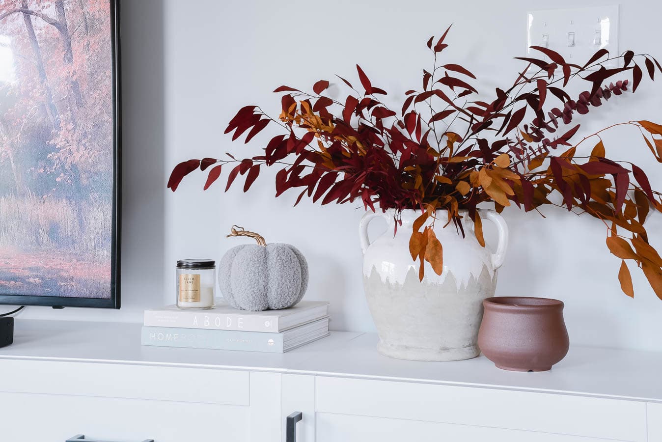 A white cabinet decorated with a white vase holding red and orange autumn leaves, a small gray fabric pumpkin, stacked books, a lit candle, and a small brown pot provides cozy fall home decor updates. A portion of a television screen displaying an autumn scene is visible to the left.