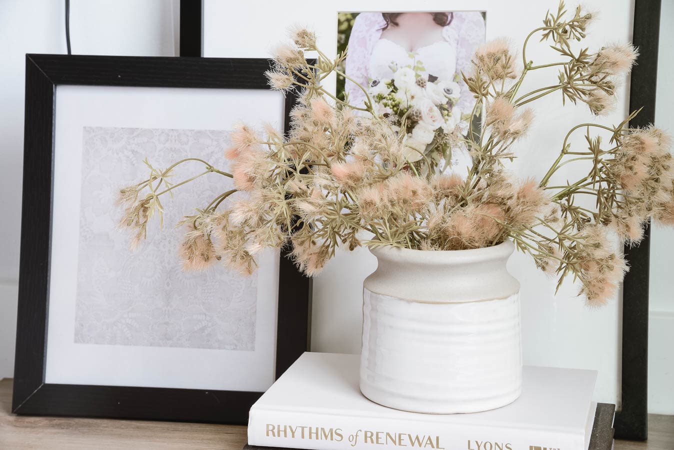 A white ceramic vase containing a bouquet of dried, pinkish flowers sits atop a stack of books. Behind the vase are two framed items: one with a patterned image and another partially visible photo of a person in a wedding dress holding a bouquet. These easy and affordable swaps can warm up your home effortlessly.