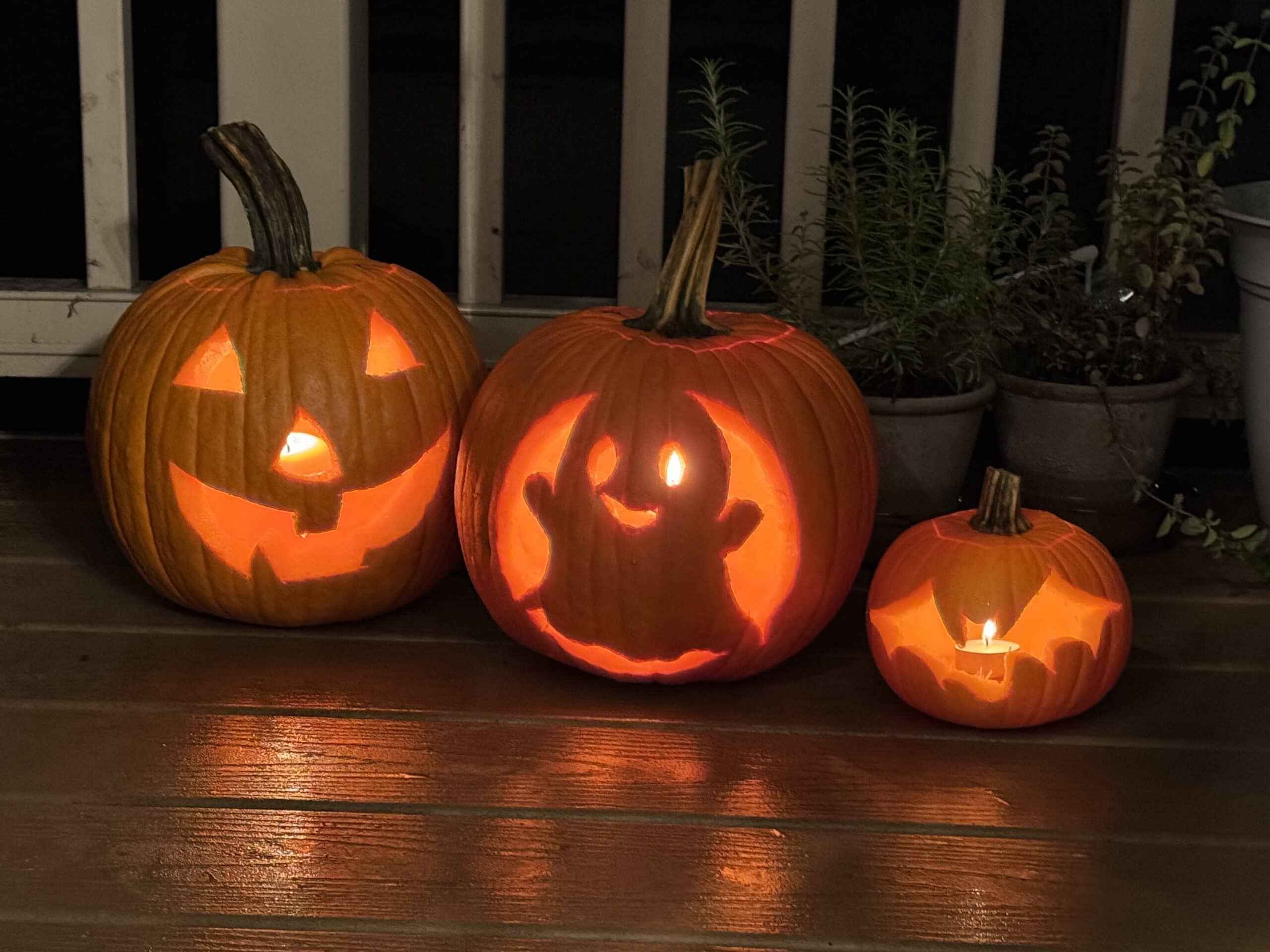 Three carved pumpkins are placed on a wooden porch. The largest pumpkin has a classic jack-o'-lantern face, the medium pumpkin displays a cute ghost using printable stencils, and the smallest pumpkin features a bat design. Each pumpkin is lit from inside, illuminating the carvings.