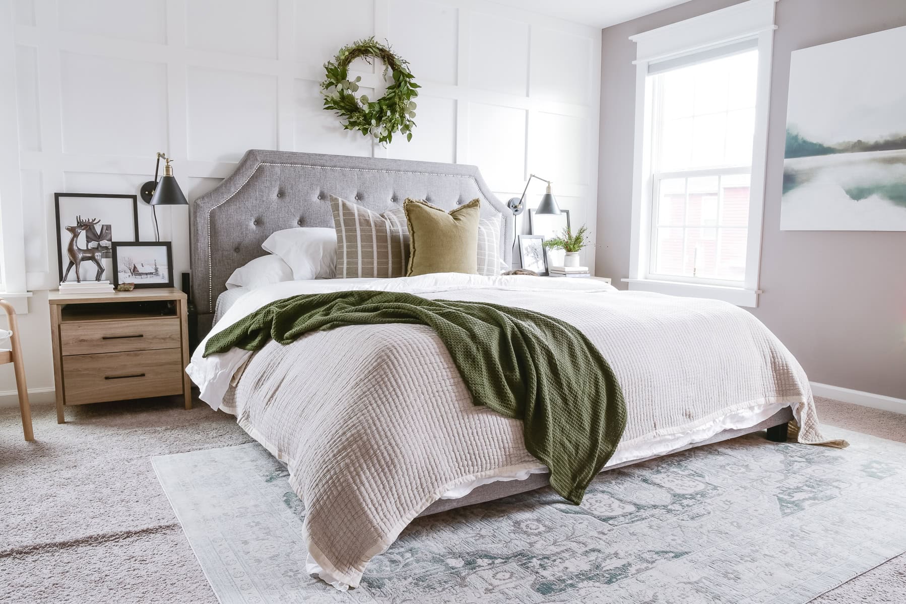 A cozy bedroom features a large bed adorned with a beige comforter and green throw blanket, embodying the essence of home decor. A wreath above the tufted headboard hints at the transition from fall to winter. Natural light spills through the window, highlighting nightstand lamps and artwork on either side.