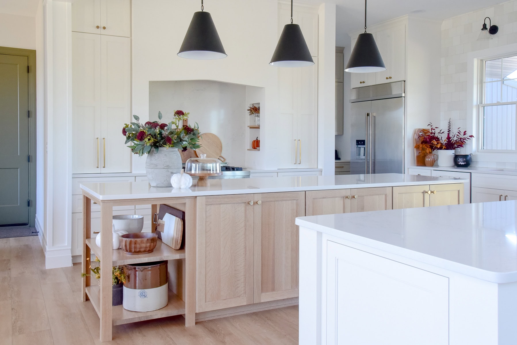 A modern and functional kitchen features white cabinets, light wood flooring, and a large island with a white countertop. Three black pendant lights hang above the island, which is decorated with vases of flowers. Stainless steel appliances are in the background, enhancing kitchen organization.