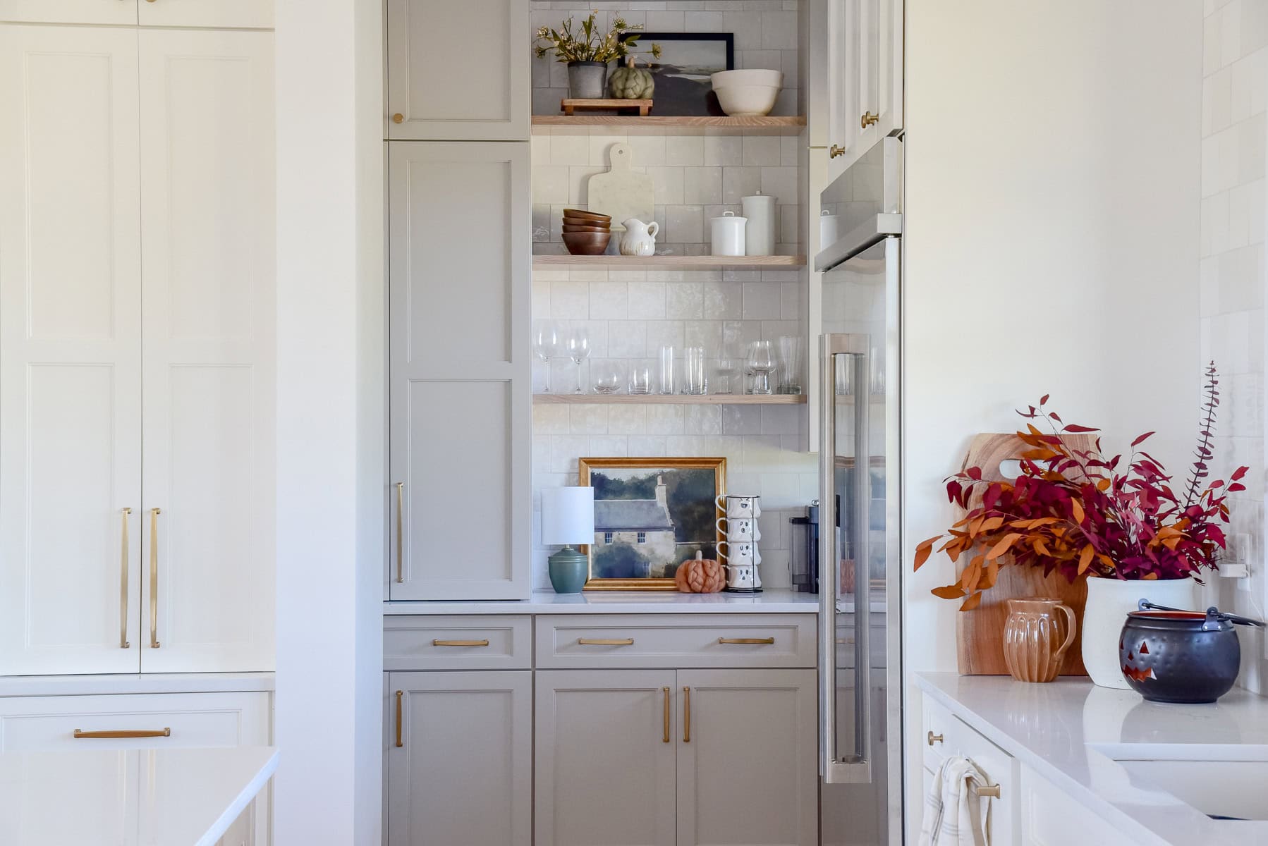 A modern, stress-free kitchen with light gray cabinets, open wooden shelves displaying glassware, framed art, and decor. A stainless steel refrigerator stands to the right, and a white countertop with flowers in a vase is perfect for preparing Thanksgiving feasts.