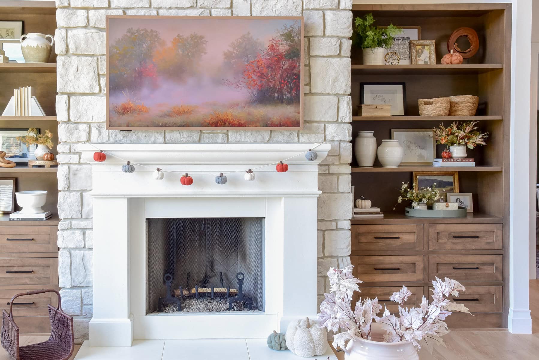 A cozy living room features a HiSense Canvas TV above a white stone fireplace adorned with a garland of small pumpkins. A landscape painting with autumn foliage complements the setting, while wooden shelves on both sides display vases, books, and decorative items.