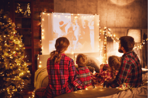 A family of four, clad in matching plaid pajamas, cozies up on a bed for a delightful Christmas Movie Night. Warm yellow string lights and festive decor illuminate the room, as the scent of holiday snacks fills the air and a Christmas tree glows beside them.