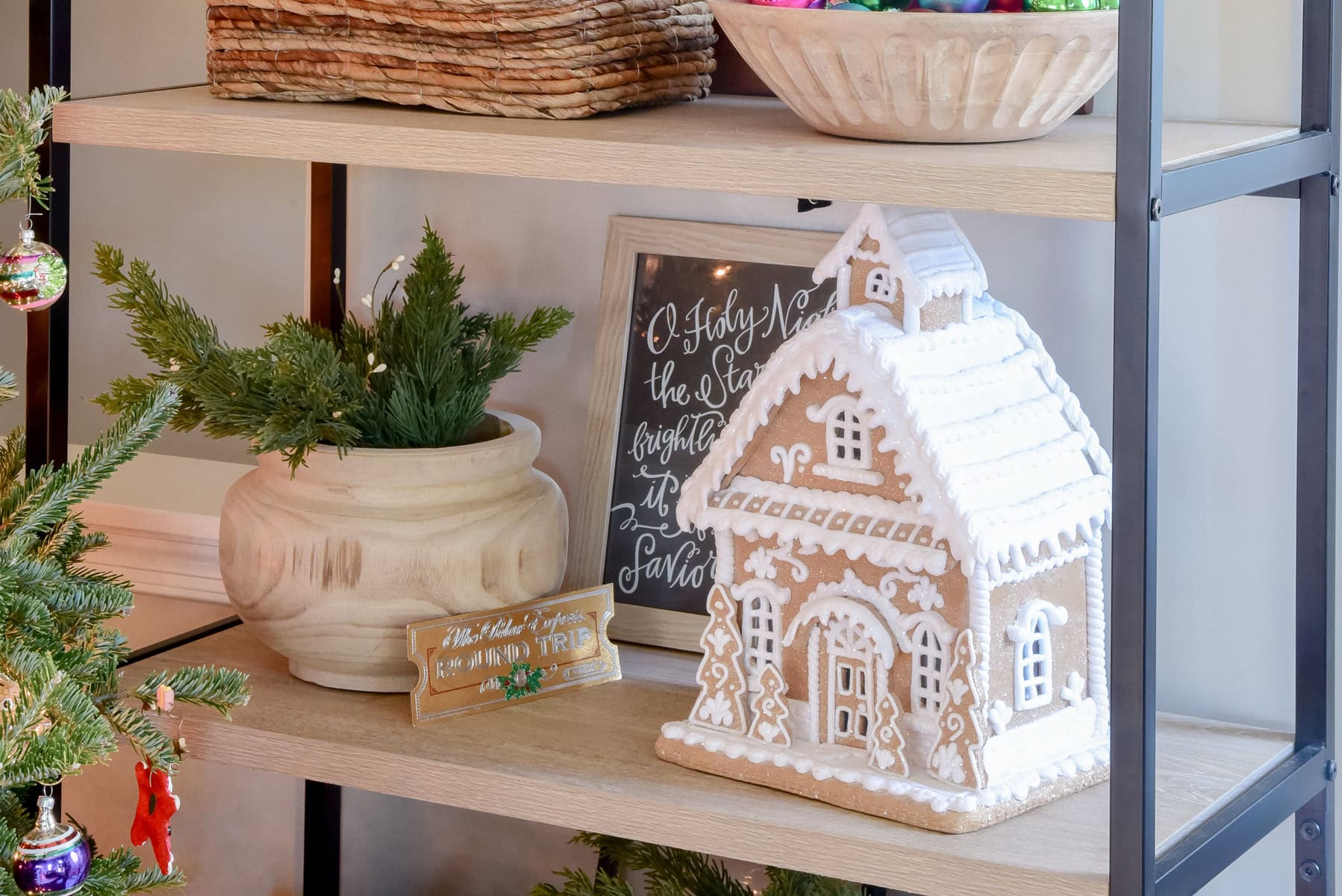 A gingerbread house with white icing details is displayed on a wooden shelf as part of this year's Christmas decor. Nearby, there's a potted evergreen plant and a framed chalkboard sign with decorative writing. A wicker basket and colorful ornaments are also visible.