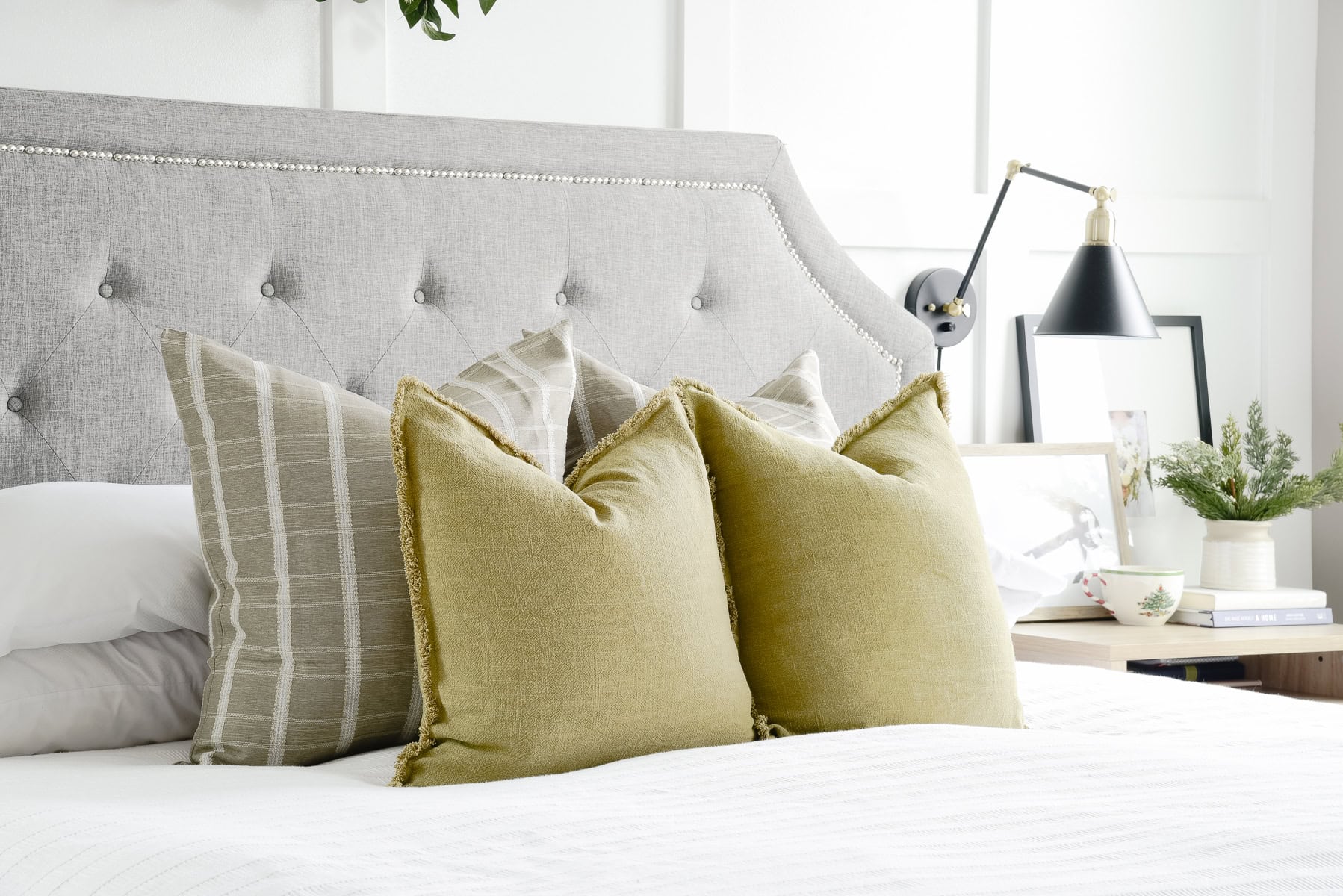 A neatly made bed with a tufted gray headboard is adorned with two green throw pillows and two plaid pillows. To impress holiday guests, the small bedside table holds a black lamp, a plant, and picture frames, creating a cozy and stylish home setting.