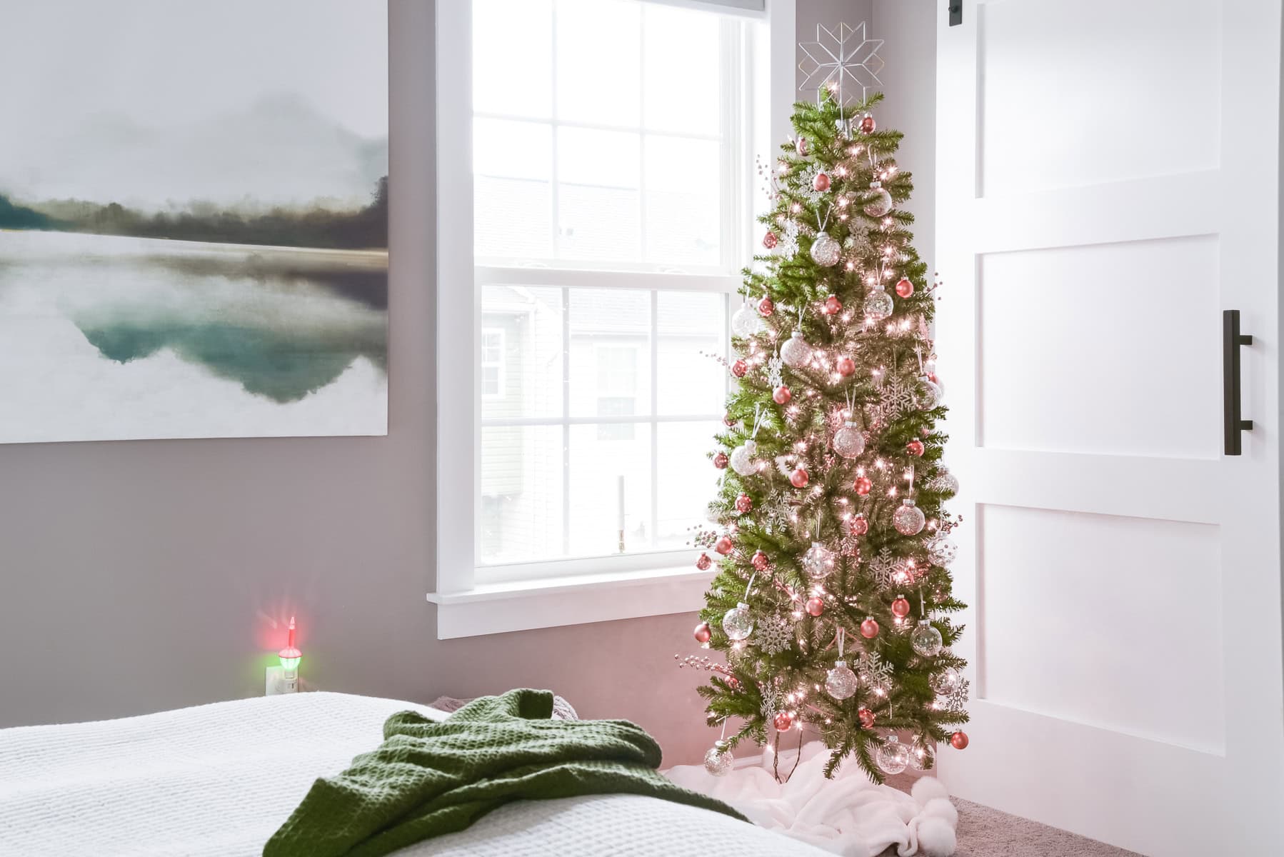 A decorated Christmas tree with twinkling lights and ornaments stands by a window in a bright, modern room. Amidst the festive charm, there's a white bed with a green blanket in the foreground, while abstract wall art adorns the gray wall, embodying holiday joy without clutter.