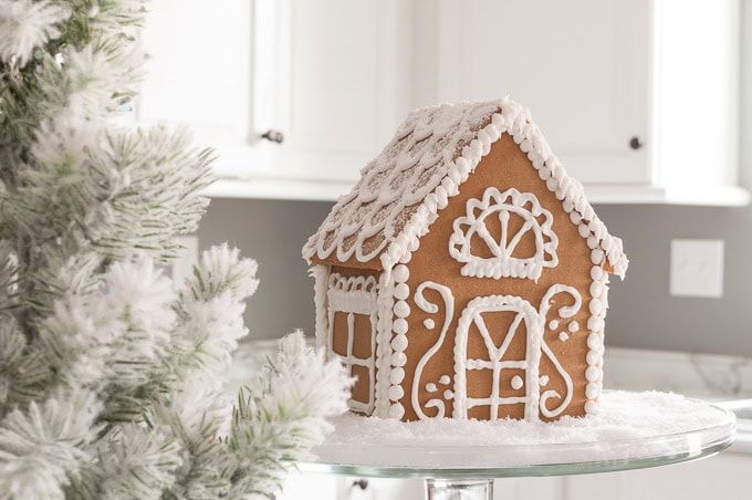 A gingerbread house adorned with white icing and candy sits on a glass tray, surrounded by snow-like frosting. In the foreground, a snow-dusted evergreen branch adds to the festive charm of organizing your kitchen for Christmas amid the bright, white background.