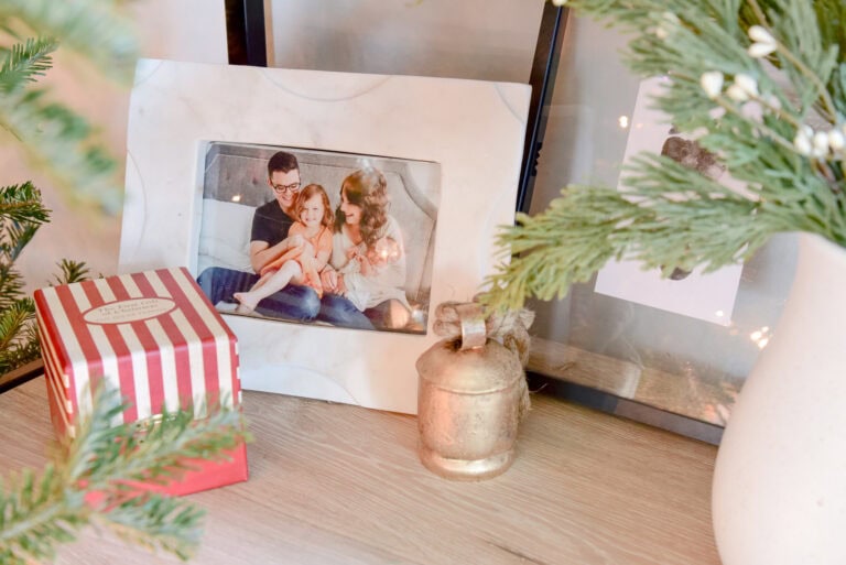 A framed family photo of three people is surrounded by festive Christmas decorations, including a striped gift box and a metallic bell ornament. Pine branches add a touch of greenery to the scene, creating a stress-free holiday ambiance.