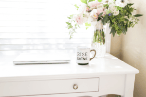 A white desk features a closed laptop and a mug that reads "Rise & Shine." Beside it, a vase brimming with pink and white flowers sits in perfect harmony. Sunlight streams through the window blinds, illustrating the benefits of decluttering and creating serene simplicity.